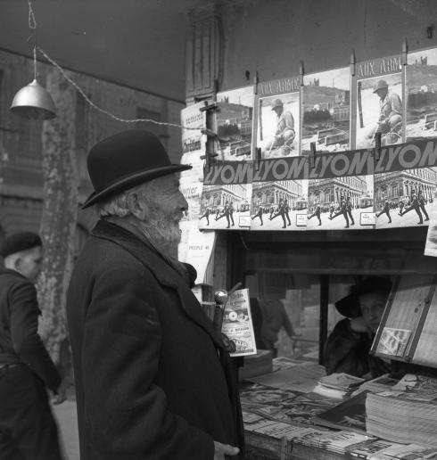 Un homme regarde un kiosque de journaux, où apparait le recueil de photographie d'Emile Rougé "Lyon sous la botte"